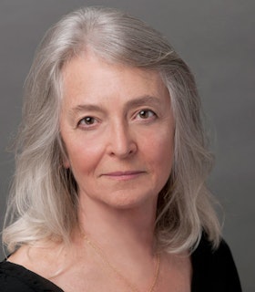 Portrait of Joan La Barbara staring at the camera with shoulder length silver hair and a black top against a gray backgorund.