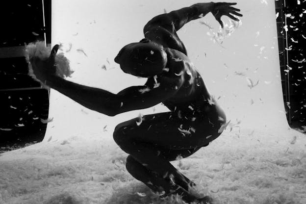 A black and white image of a performer kneeling with one extended arm forwards and the other backwrads holding feathers which surround them on the floor.