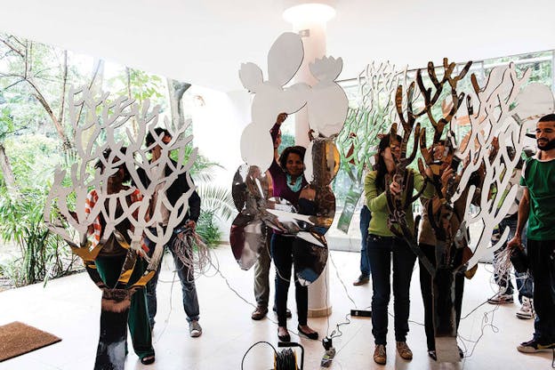 Persons hold up mirrors shaped like cacti in a bright white room wallpapered with palm plants and thin tree trunks.