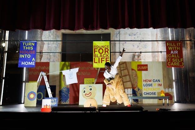 Nile Harris stands in the middle of a backlit stage, bent forward towards the audience with brightly colored posters, stage sets, and other props includeing a ladder, a rolling trolley, a portrait of a gingerbread man, and a white graphic t-shirt, arranged behind him. His feet are apart, torso pitched forward, and his left hand is raised making a peace sign above him. He is wearing a gingerbread cookie costume with his upper half removed from the sleeves, a white longsleeved t-shirt is underneath.