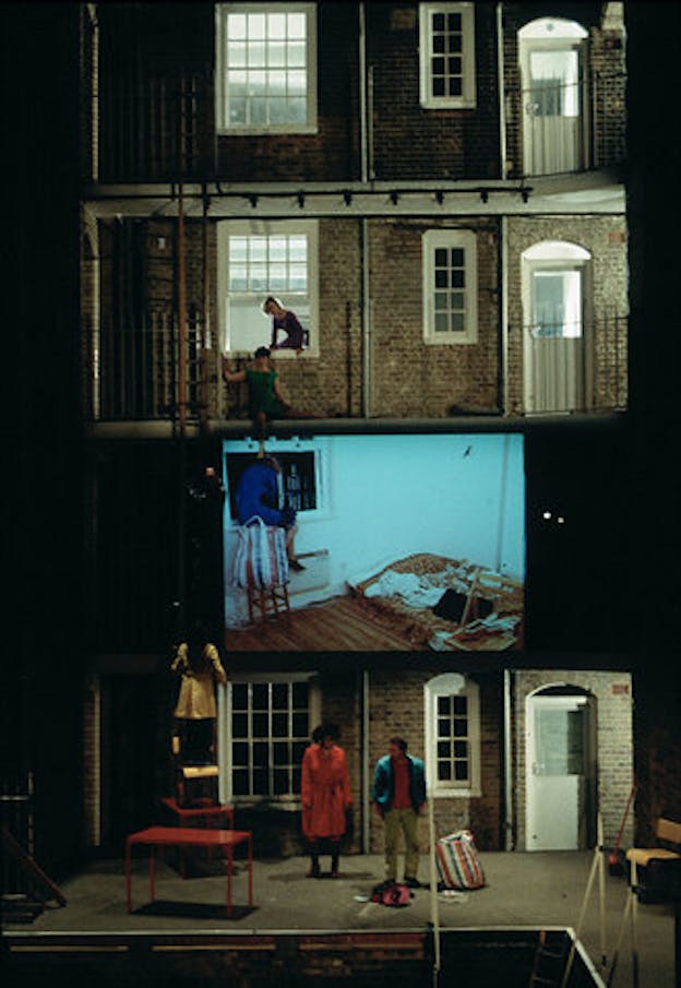 The facade of a staged building contains in its middle a screen projection of a figure looking outside a window. Three performers stand on the front while two others are situated on a window on top of the projection.