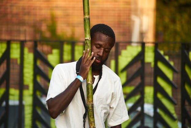 A photograph of JJJJJerome Ellis performing, holding a natural reed close to him, closing his eyes and looking downwards. He stands in front of a brick wall and a black fence and wears a textured white shirt.  