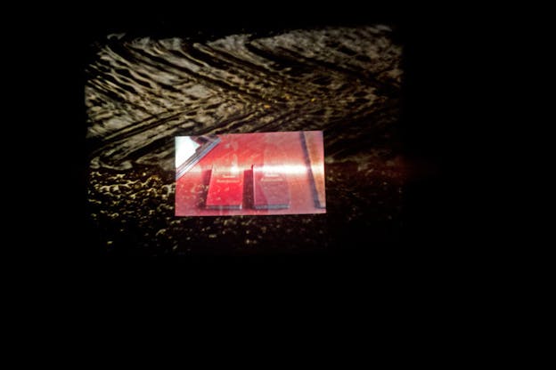 Small rectangle projecting two side-by-side books washed in stoplight red light with a white flare across the middle, superimposed on a larger rectangle projecting what appears to be a black and white close up of a stream on gravel, suspended in black space.