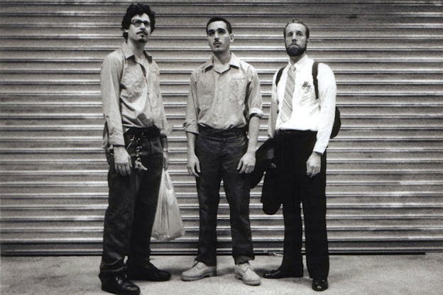 A black and white image of three performers who stand in front of a closed metal gate and look dejectedly in front of them. The two performers on the left, wear matching grey shirts and dark jeans. The performer on the right wears a white shirt, a striped tie, and dark pants. 