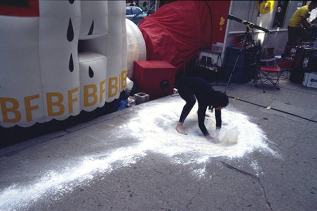 A performer wearing all black spreads white dust around a city sidewalk. Behind them, there is an inflated white object with the letters B and F printed on it. 