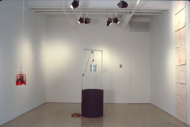 A makeshift well with a maroon bottom and a silver bucket hanging from it stands in the middle of a room with wooden floorboards. A red cage hanging from the ceiling is situated to its left. 