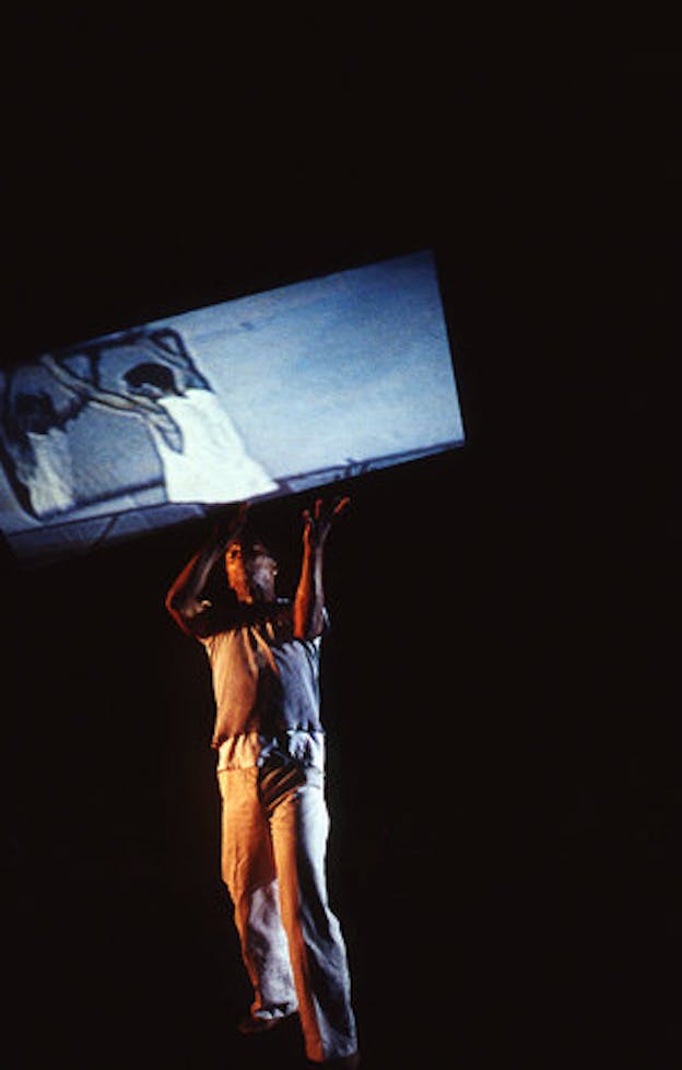 A performer wearing brown pants and a brown shirt lunges forward slightly in a dark space. They hold a screen above their head which projects a black and white image of people in white tank tops facing each other and holding their arms up above their head.