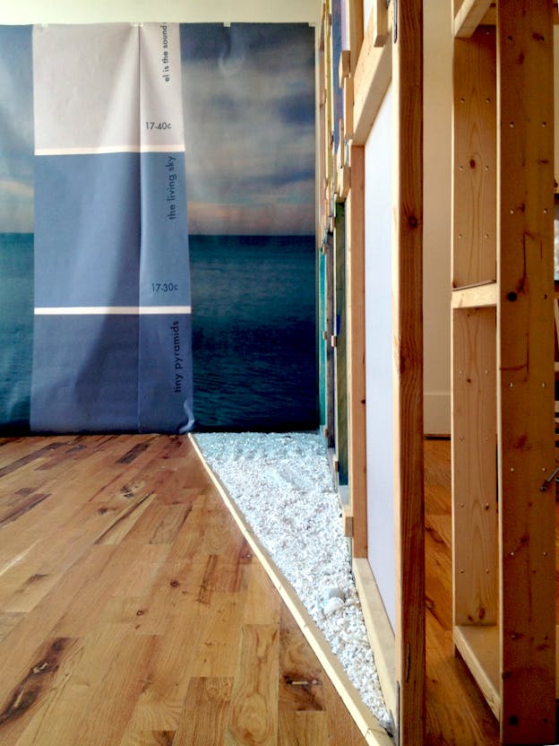 Installation view of a wall papered with an image of a twilight-blue ocean horizon separated by a tri-color blue sheet displaying the words 