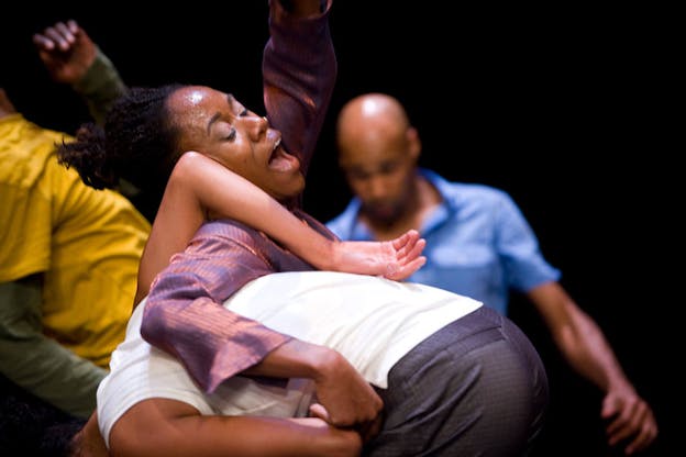 Close-up of a performer closing their eyes and opening their mouth while raising one arm and holding on to the bent back of another performer. Blurred behind them, two performers dance.