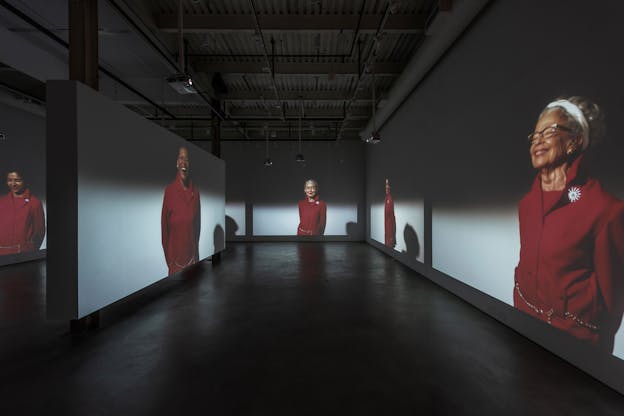 Installation view of figures clad in red smiling projected on the walls of a makeshift hallway. 
