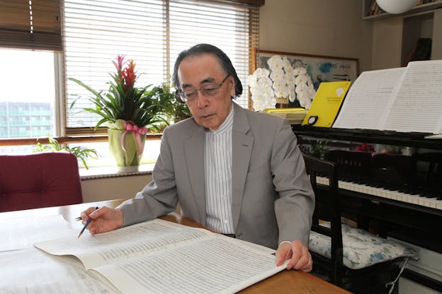 A person in glasses and a gray suit with a striped button up underneath seated with their back to a piano on a wooden table, looks down on music sheets scattered around the table.