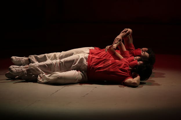 Three performers lie in a row on their left sides, one in front of the other. Each wears a red mask, red shirt, white pants, and white shoes. Their left arms are behind their heads and their torsos turn slightly as their right arm curves over their body.