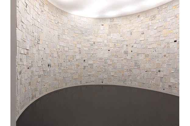 A gallery room with a gray floor and a circular shapes wall full of papers with writing on them.