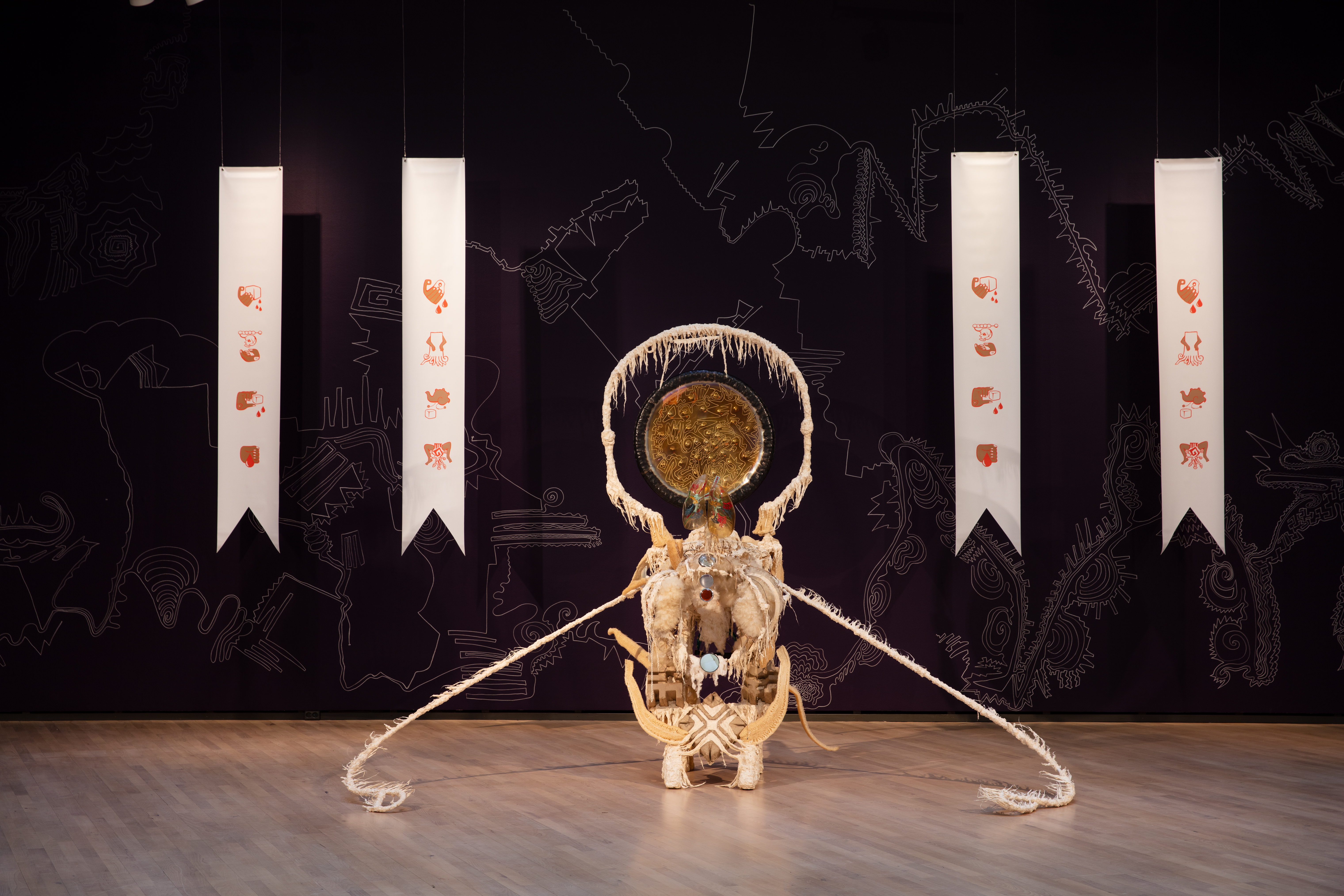 A gallery space with wooden floors and black walls with white ornamental, almost cartographic line drawings. Two white banners hang vertically on either side, framing an off white sculpture centered in the frame. They are each decorated with four minimal icon illustrations. The sculpture is composed of off white organic materials that form a small pedestal, decorated with cotton, loofah, and carved wood. One long ragged strand extends out from each side and extends to the floor. On top of the pedestal sits a hoop made from the same textured, fringed material. Centered and hanging inside the hoop is a golden metallic plate with linear designs carved in its surface along with dents from bullets. At the base of the hoop on top of the pedestal is a transparent model of a pair of lungs, painted with red flowers.