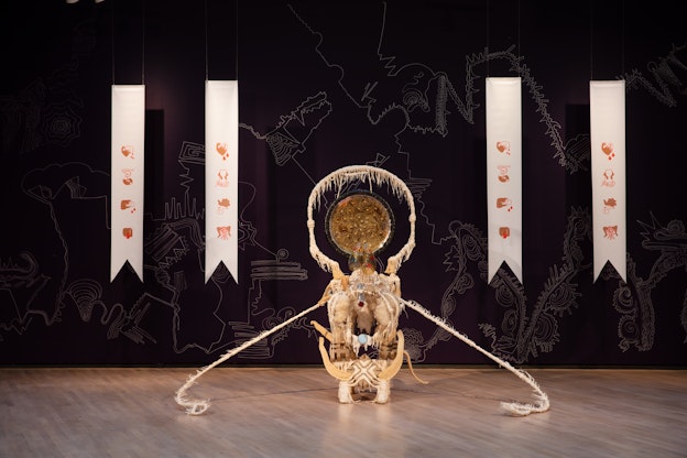 A gallery space with wooden floors and black walls with white ornamental, almost cartographic line drawings. Two white banners hang vertically on either side, framing an off white sculpture centered in the frame. They are each decorated with four minimal icon illustrations. The sculpture is composed of off white organic materials that form a small pedestal, decorated with cotton, loofah, and carved wood. One long ragged strand extends out from each side and extends to the floor. On top of the pedestal sits a hoop made from the same textured, fringed material. Centered and hanging inside the hoop is a golden metallic plate with linear designs carved in its surface along with dents from bullets. At the base of the hoop on top of the pedestal is a transparent model of a pair of lungs, painted with red flowers.