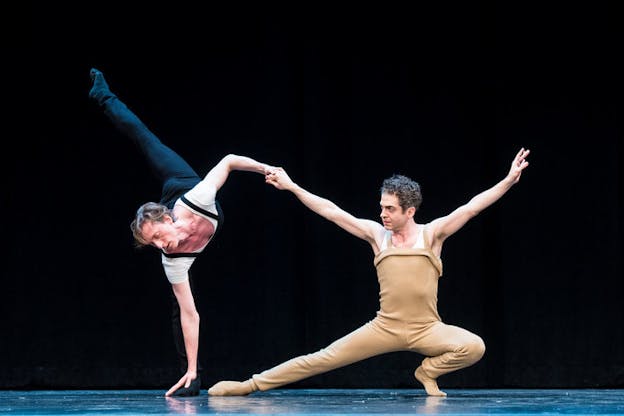A performer dressed in beige crouches down with one leg extended to the side and both arms upwards. They hold with one hand another performer to their right, who lifts one leg backwards and touches with their left hand their left foot. 