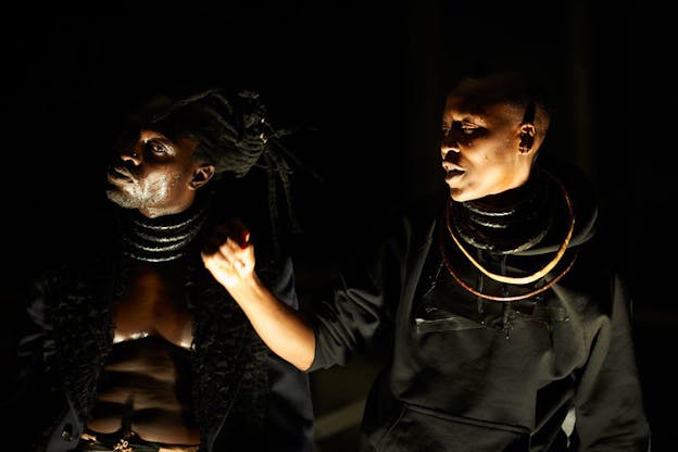 Two performers dressed in black clothing and ring necklaces adorning their necks have their shadowed faces sideways while their bodies face forward.