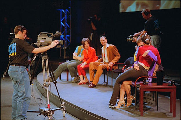 Several performers sit on stage. Two performers in the foreground are locked in an embrace which a camera man records. On the left, three performers sit and watch appearing aghast. Another camera man records them.