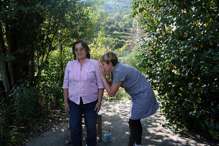 A figure stands straight en face the camera while another next to her stands side ways with their back hunched and their hands covering their mouth that is situated on the other's upper arm.