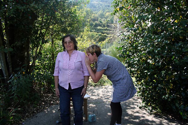 A figure stands straight en face the camera while another next to her stands side ways with their back hunched and their hands covering their mouth that is situated on the other's upper arm.