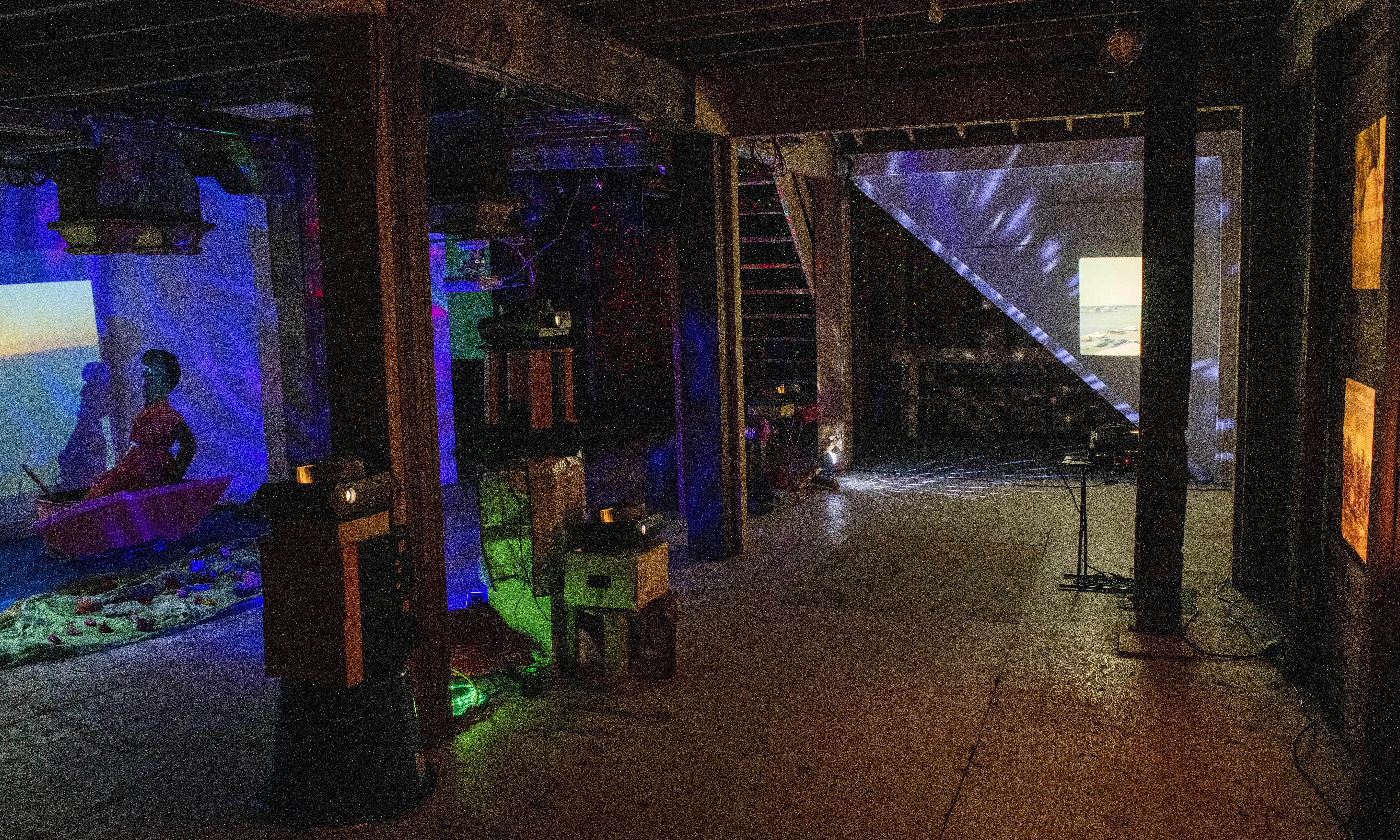 An room-sized installation in a dimly-lit basement featuring a life-size sculpture of a person inside of a boat and multiple projectors scattering points of light and projections across the walls.