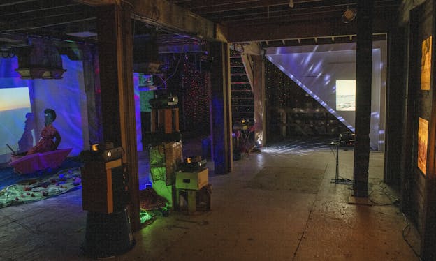 An room-sized installation in a dimly-lit basement featuring a life-size sculpture of a person inside of a boat and multiple projectors scattering points of light and projections across the walls.