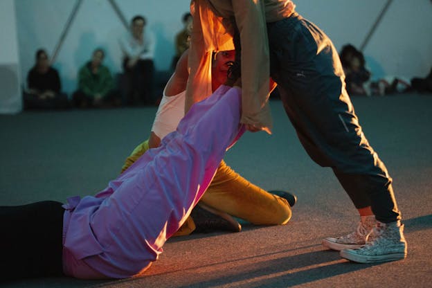 In a carpeted space, one performer lays on the ground with their feet up, supporting the body of another performer who leans on them. Behind them, another performer sits on the ground with their body angled toward them. The performers wear vibrant, loose-fitting pants and are lit by a warm spotlight. An audience sits on the ground in the blurred background and watches.