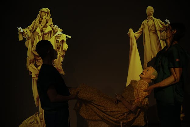 devynn emory and Joseph Pierce are silhouetted in the foreground of the image, carrying a medical mannequin wearing a hospital gown between them. In the background are two sculptures of figures with outstretched arms draped in yellow cloth.