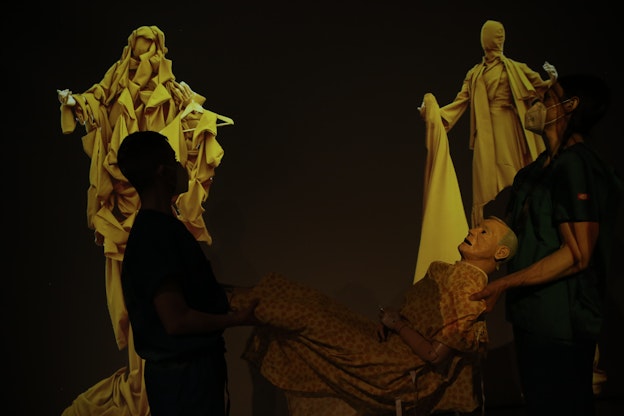 devynn emory and Joseph Pierce are silhouetted in the foreground of the image, carrying a medical mannequin wearing a hospital gown between them. In the background are two sculptures of figures with outstretched arms draped in yellow cloth.