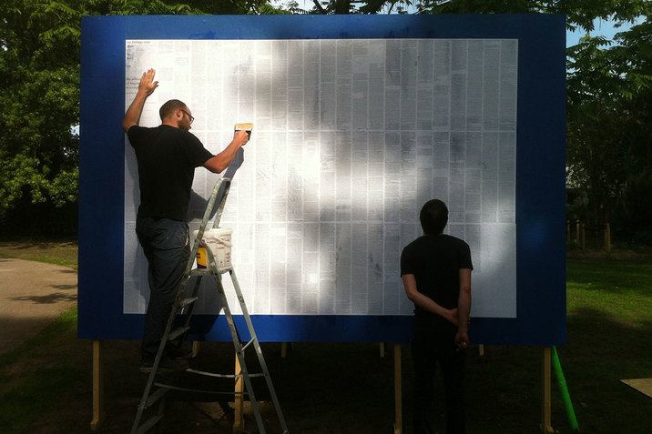 A person dressed in a gray suit dances and twists their body in the center of a black stage with gray floors while a person standing in the back left smiles and watches. 