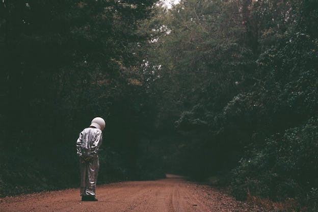 Wide-angle shot of a back-facing person dressed in a space suit and helmet standing and slightly bending their head in a dirt road lined with dark trees. 