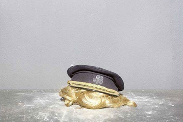 Still-life of a blonde wig attached to a black officer cap on a metal gray table set against a light gray wall. 