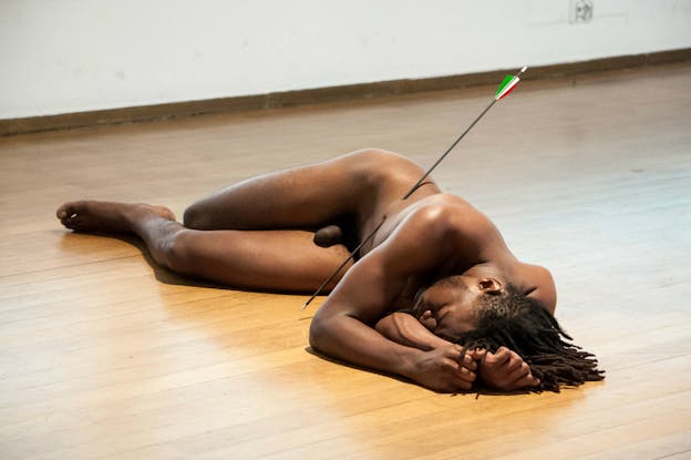 A performance still of Carlos Martiel laying on his side on a wooden floor, nude, with an arrow piercing his stomach. His eyes are closed and his arms are up by his head.  