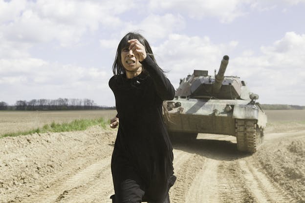 A figure dressed in black runs in a soil plain being chased by a tank.