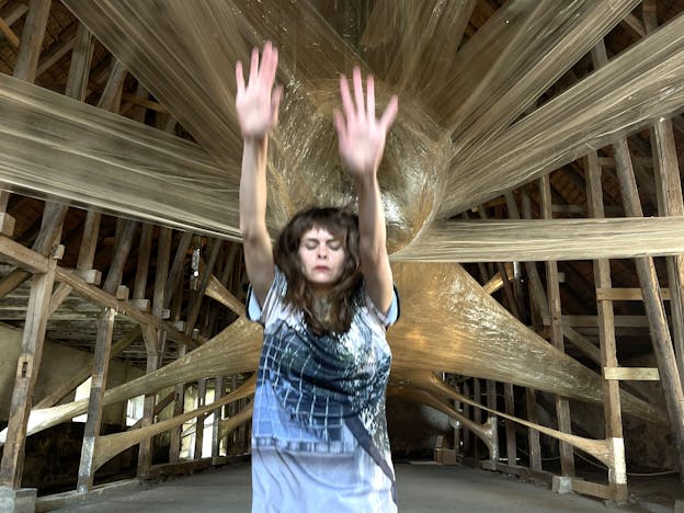 Joanna Kotze performs inside a large wooden building, the sloped ceilings and wooden support frame of the building are visible in the background. A large tan cocoon-like sculpture is mounted behind her in the middle of the space. Her body is blurred mid movement, she faces the camera with her eyes closed as she jumps straight up, her arms reaching up, palms facing the camera. She is wearing a long white t-shirt printed with the image of a grid fence.