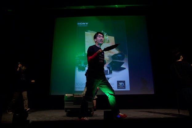 Low angled view of a performer onstage holding out a closed umbrella, their dimly lit body smattered in green and red light from a screen projecting a Sony advertisement.