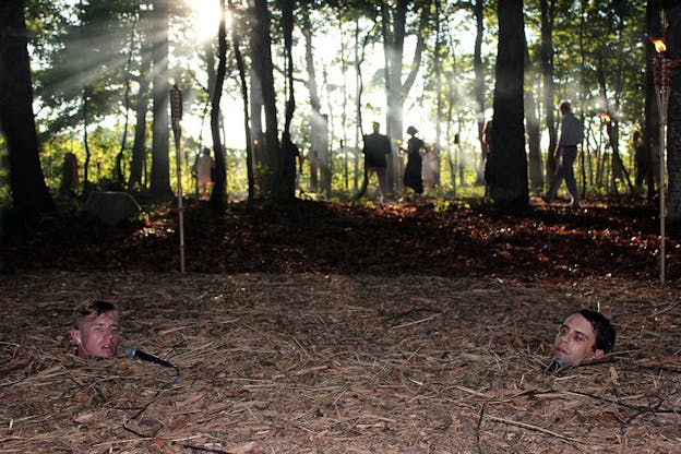 Two heads with microphones placed at their chins poke out of a forest floor of dried pineneedles and sticks while behind them figures walk through a sun-dappled forest. 