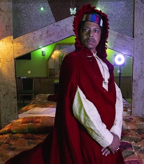 Azikiwe Mohammed stands sideways with his hands clasped in front of him looking directly into the camera. Behind him is a bed covered with a quilted cover and a mirrored headboard shaped like a pentagon. He wears a white button-down shirt and red hooded cloak.