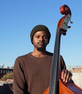 Luke Stewart stands on a rooftop with his upright bass looking calmly into the camera. He wears a stocking cap and long-sleeved brown sweater. Blue sky and a city skyline are in the background.