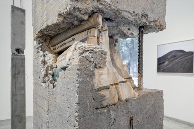 A close up image of one of the concrete pillars partially ripped off in the middle. The fracture reveals the interior of the pillar that is made of stacks of books. One of the book spines has a legible book title in black that reads: 