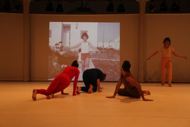 Performers with their backs towards the viewer lay on the ground on various poses as they watch in front of them on the wall the projection of a child with its arms slightly raised. Another performer on the side of the projections en face the viewer holds a similar pose.