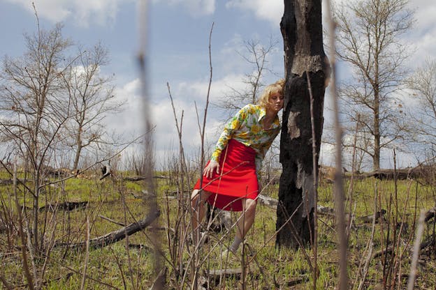 Person in a bright red skirt and yellow floral blouse closes their eyes and leans against a burnt tree in a freshly green meadow of thin budding trees. 