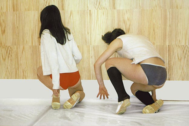 Two performers wearing white tops, brightly colored underpants, and white shoes crouch on a white floor in front of a wooden wall lwith their backs turned to the camera. 