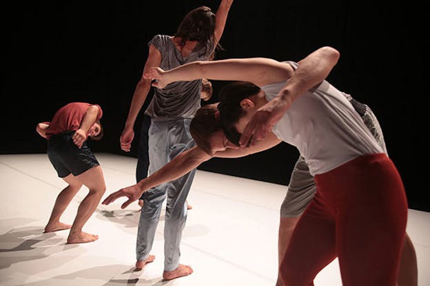 Four performers on a white stage. The two closest to the viewer move their bodies forward, their arms opens. The one behind them, stands with one extended upwards. The fourth performer, farthest away from the viewer crouches their upper body. 