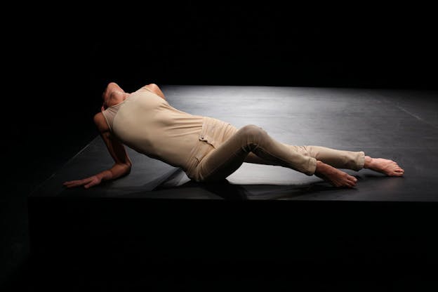 A performance still of Maria Hassabi laying on a black stage, wearing cream colored jeans and a cream colored tank top. She rests on her elbows and bends back, putting her chin towards the air.