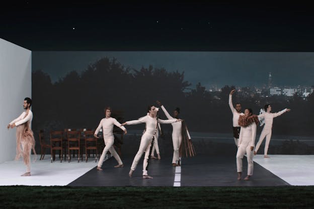  Performers dressed in white unitards and varying brown accompaniments pose on a stage flanked in the front with green grass, a wooden dining table, and on the back wall an image of silhouetted trees and a distant city skyline lit up at night.