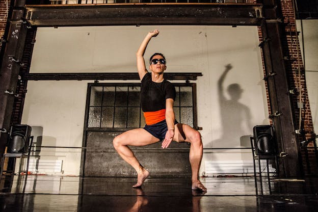 A performer dressed in black sungalsses, a black shirt, orange waist band and blue shorts squats down with their tippy toes and lifts one hand up as the other is kept in the front. 