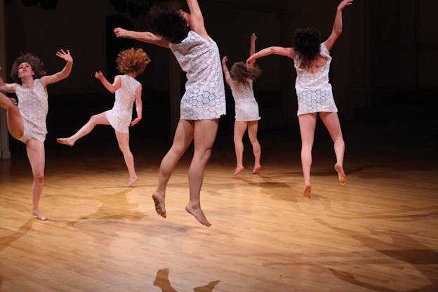 Performers in white short dressed with flower cut details jump on a wooden floor.