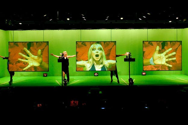 Three performers stand on a green hued stage. The performer in the middle has long blonde hair and wears a black dress. They face a camera, wearing an aghast expression on their face and hold out both of their arms at shoulder height. Their face is projected onto a screen to the left of them. Two more screens on either side of the first screen show the projection of open hands. A performer stands behind each of these screens, reaching their hand in front of a camera and concealing the rest of their bodies behind the projector screens.