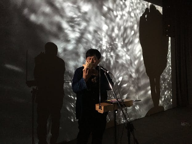 Koichi Makigami performs in front of a projected abstract silver background image . He faces the camera and stands before a theremin with a tablet and a microphone, blowing into a large conch shell. He wears a dark button up shirt.
